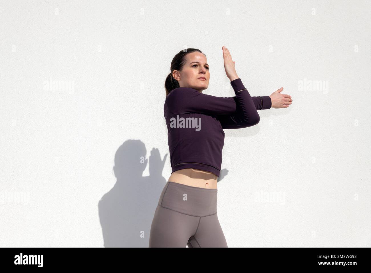 Fit sportliche Frau, die sich draußen an einer weißen Wand aufwärmen und sich die Arme dehnen lässt. Stockfoto
