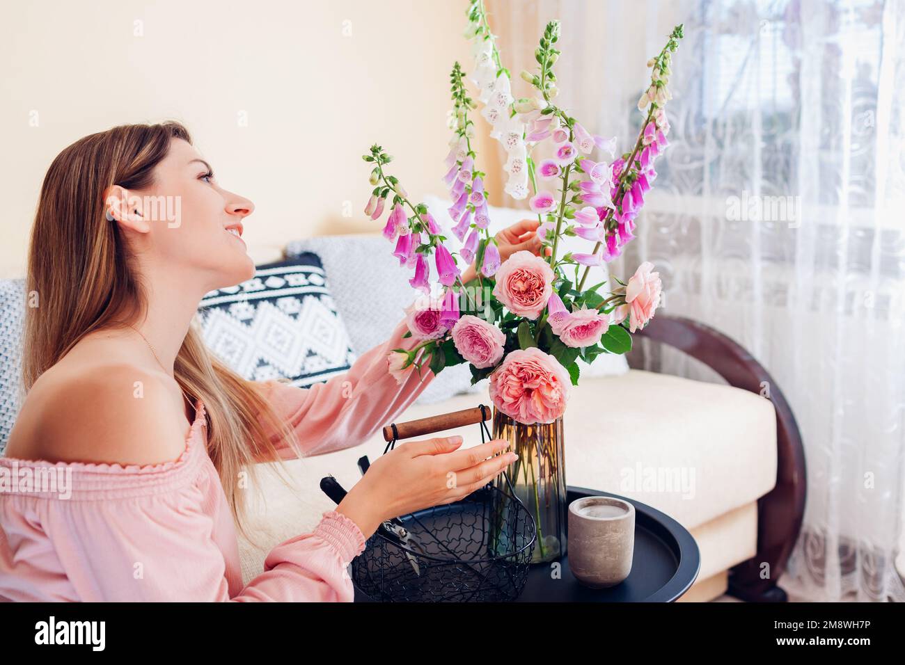 Frau macht Rosenstrauß und Handschuhblumen in Vase auf dem Tisch zu Hause. Einrichtung im Sommer. Frische pinkfarbene orangefarbene Blüten im Arrangement Stockfoto