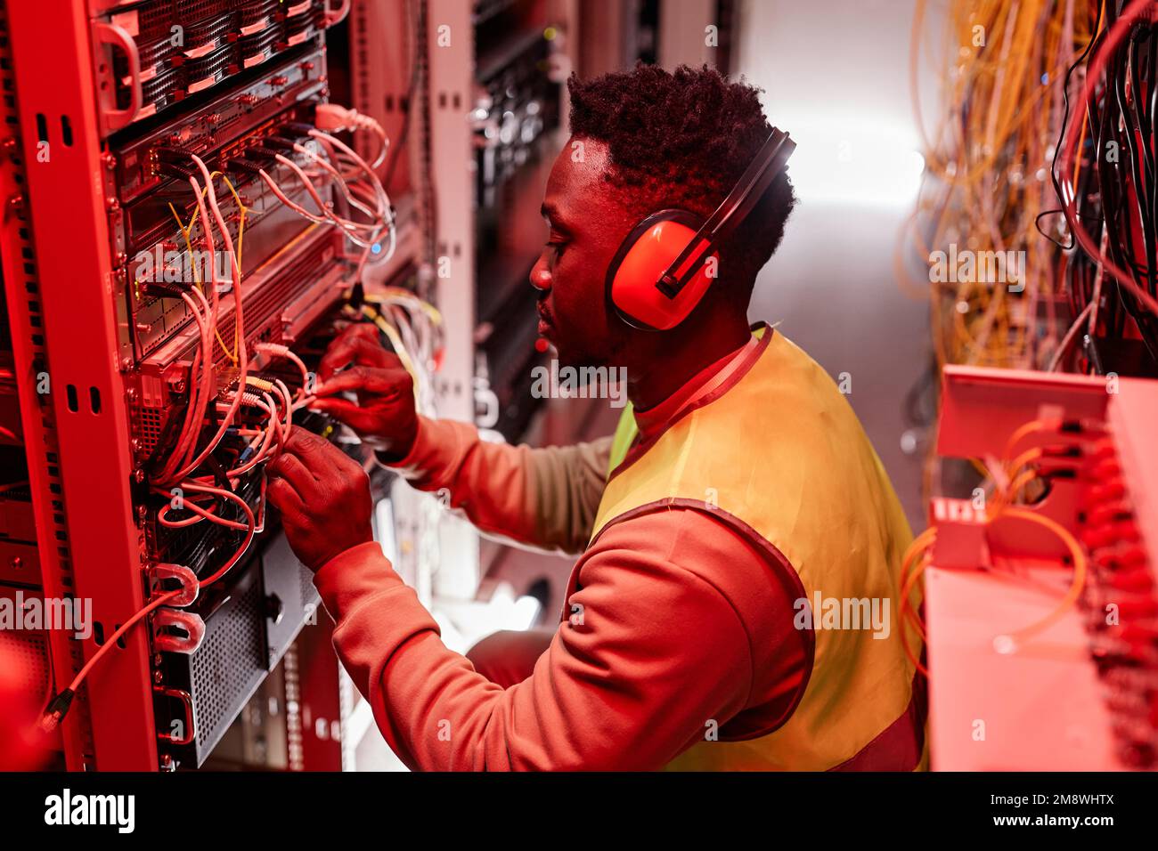 Seitliches Porträt eines Technikers, der das Netzwerk im Serverraum einrichtet und einen Gehörschutz trägt, der von roten Neonlichtern beleuchtet wird Stockfoto