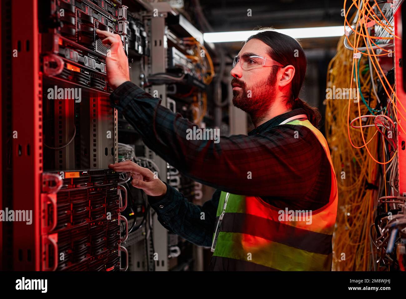 Seitenansicht eines bärtigen Netzwerktechnikers, der Server inspiziert und Wartungsarbeiten im Rechenzentrum durchführt, beleuchtet durch rotes Neonlicht Stockfoto