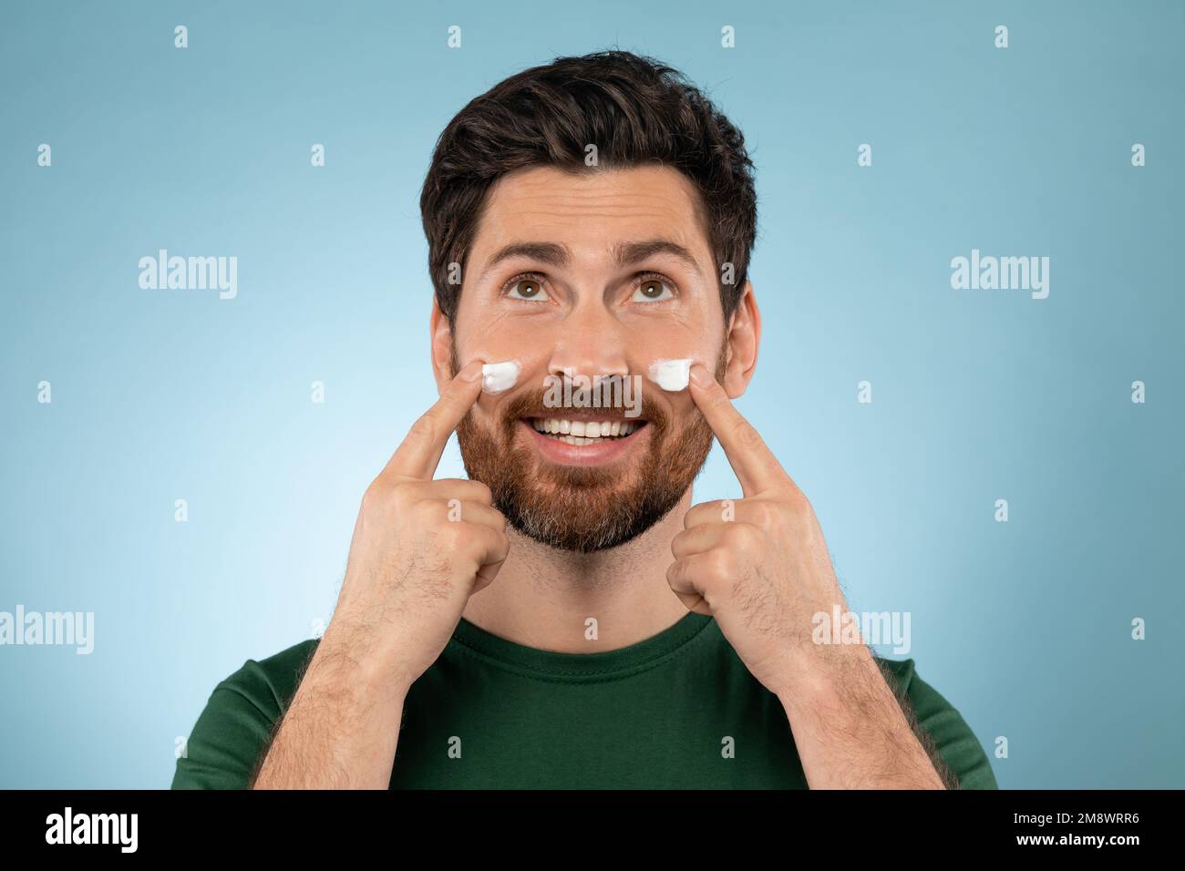 Bärtiger, gutaussehender Mann, der Gesichtscreme auf die Wangen aufträgt, auf blauem Studiohintergrund steht, in die Kamera schaut und lächelt Stockfoto