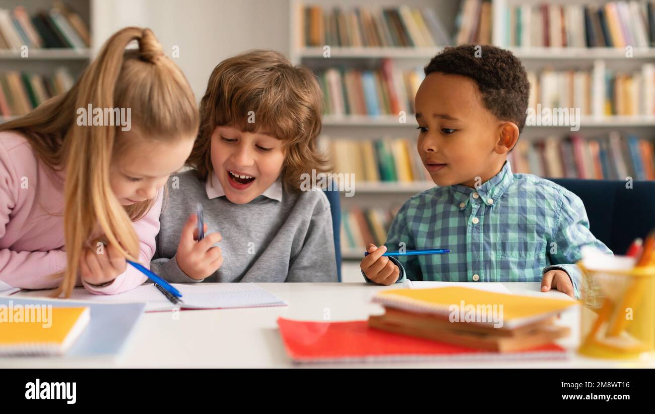 Zeit für die Schule. Multiethnische Kinder, die in Notizbüchern schreiben und lächeln, miteinander reden und lachen, am Tisch sitzen Stockfoto