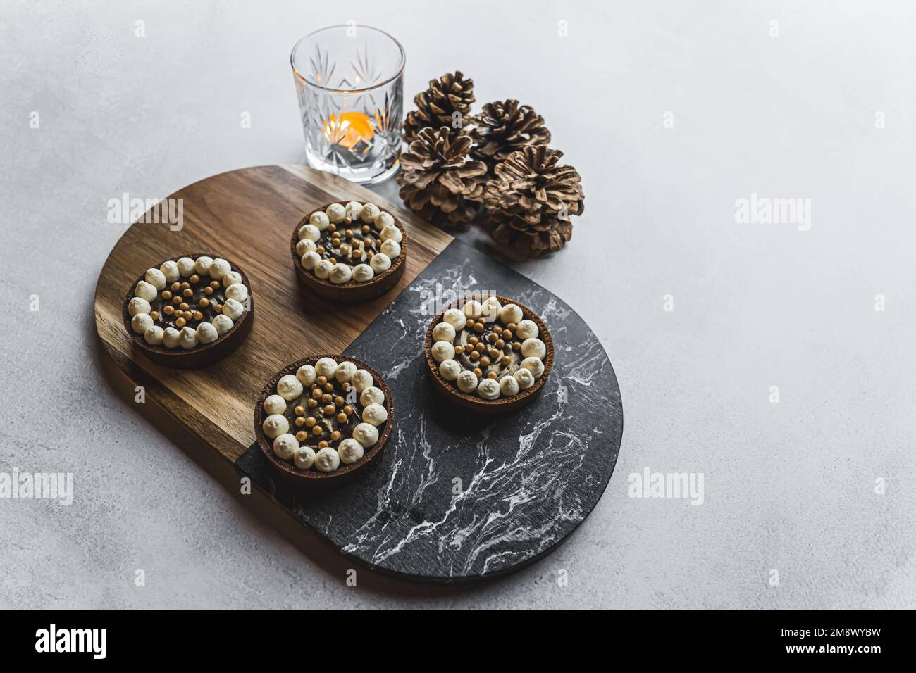 Vier Mini-Schokoladen-Desserttörtchen, serviert auf einem Holz- und Steintablett auf einem Steintisch mit Kerze und dekorativen Kiefernzapfen. Festliche Winterdesserts. Horizontale Aufnahme. Hochwertiges Foto Stockfoto