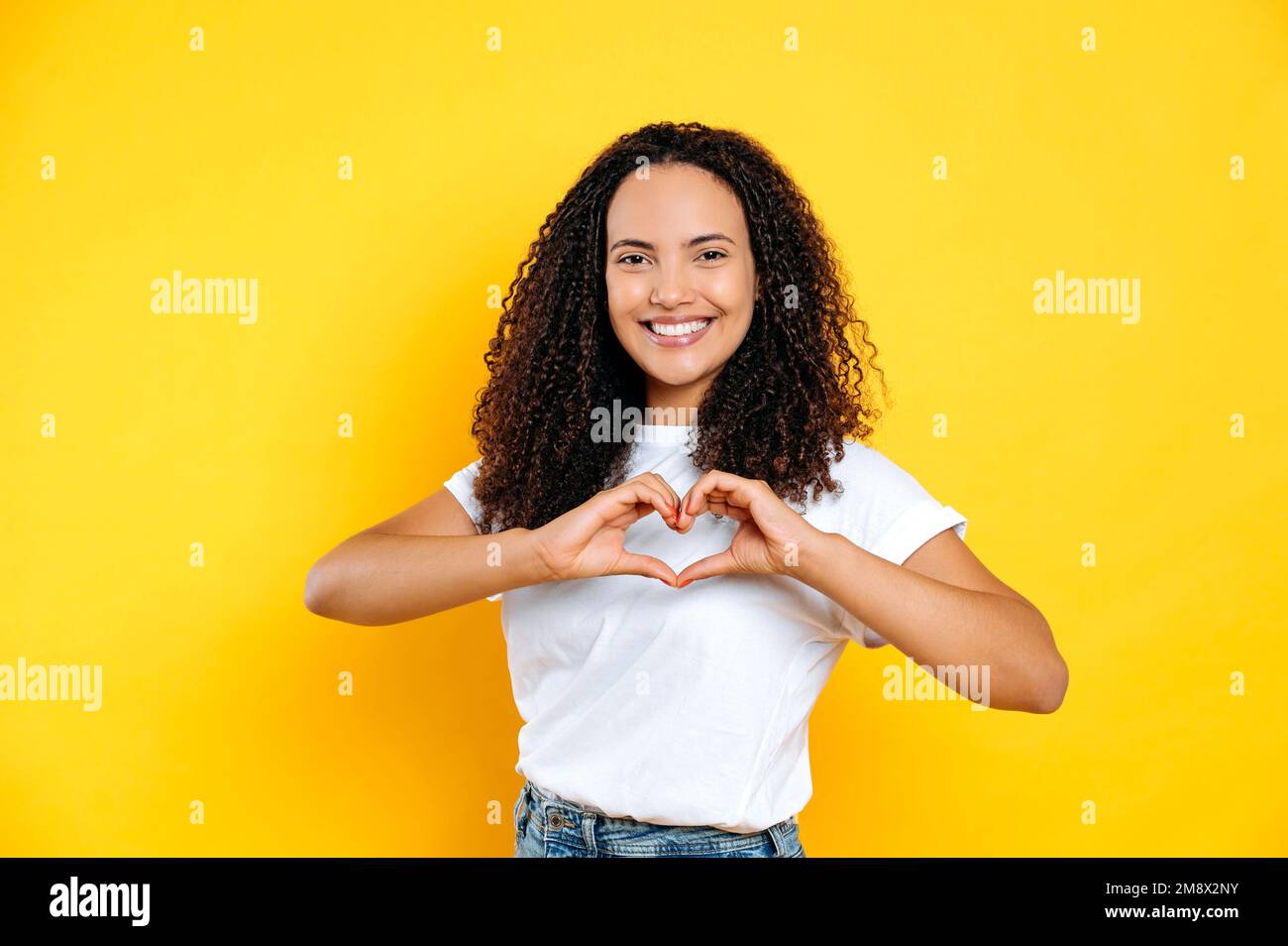 Hübscher, offener, angenehmer Latino oder junge brasilianische Frau, in einem einfachen T-Shirt, macht Herzgesten mit Händen, zeigt Liebeszeichen, steht auf isoliertem gelbem Hintergrund, schaut in die Kamera, lächelt freundlich Stockfoto