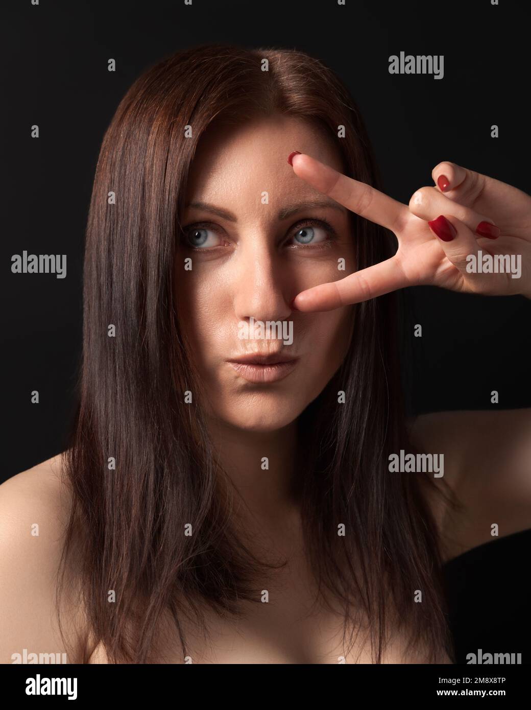 Studioporträt einer 40-jährigen Frau, die Sieg- oder Friedensgeste zeigt, mit den Fingern nahe dem Auge. Brünett mit langen Haaren und ausgestreckten Lippen, die wegschauen Stockfoto