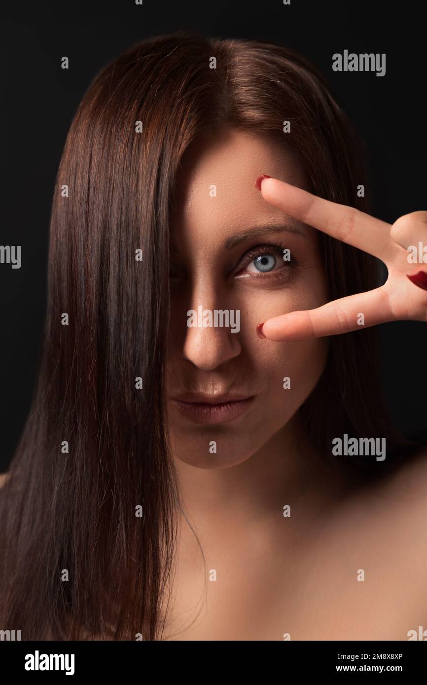 Porträt einer netten erwachsenen Dame, die ein Siegesschild nahe dem Auge zeigt und in die Kamera schaut. Reizende, brünette Frau mit langen Haaren, die ein Auge bedeckt Stockfoto