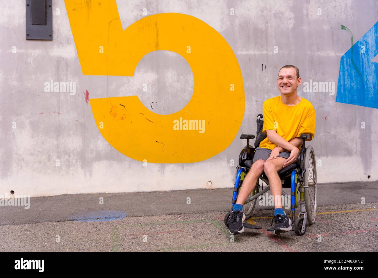 Behinderte Person in einem öffentlichen Park im Rollstuhl, trägt ein gelbes, lächelndes T-Shirt Stockfoto