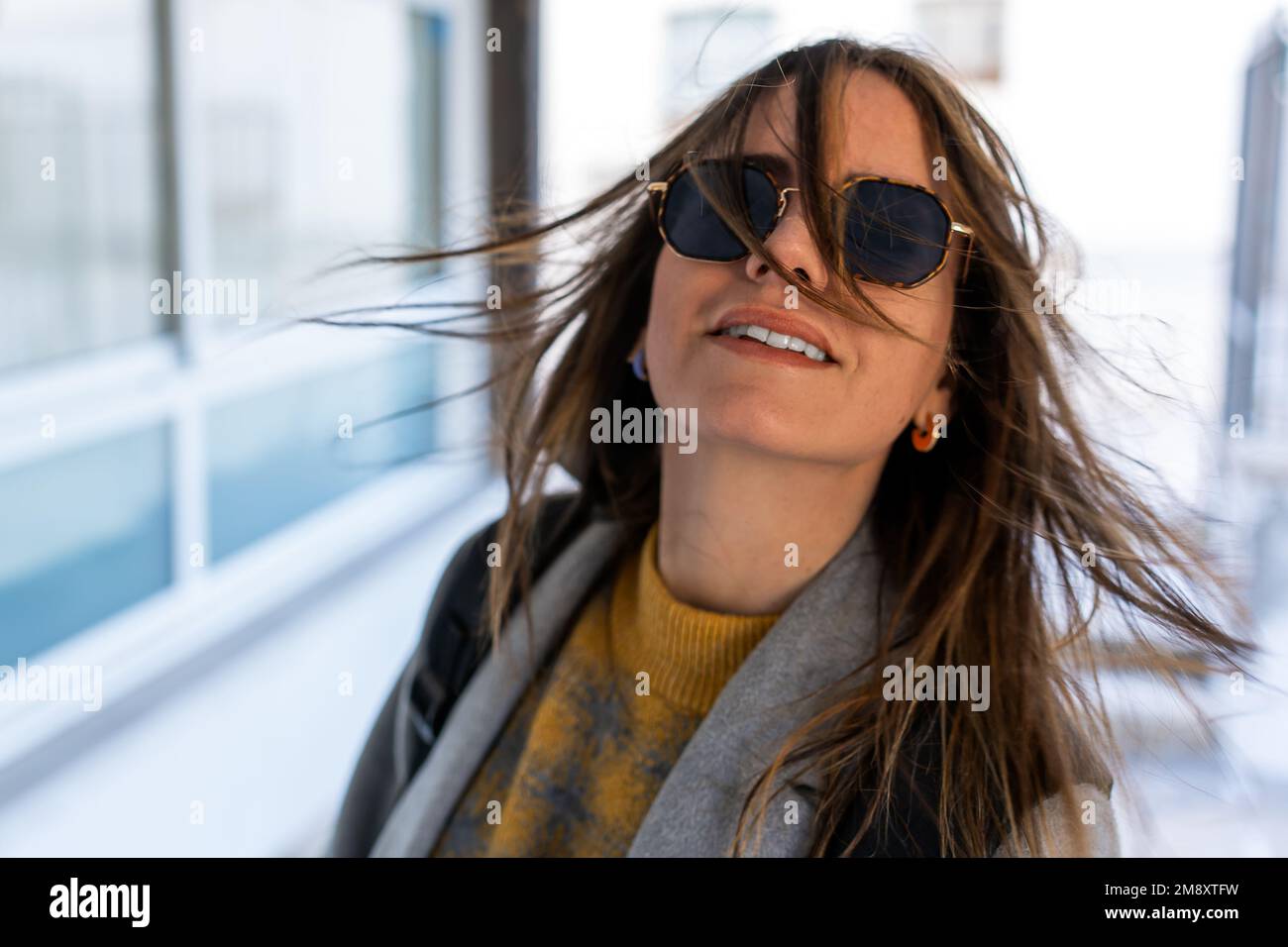 Fröhliche junge Frau in Sonnenbrille und Mantel mit fliegenden dunklen Haaren, die sich auf einem unscharfen Straßenhintergrund drehen Stockfoto