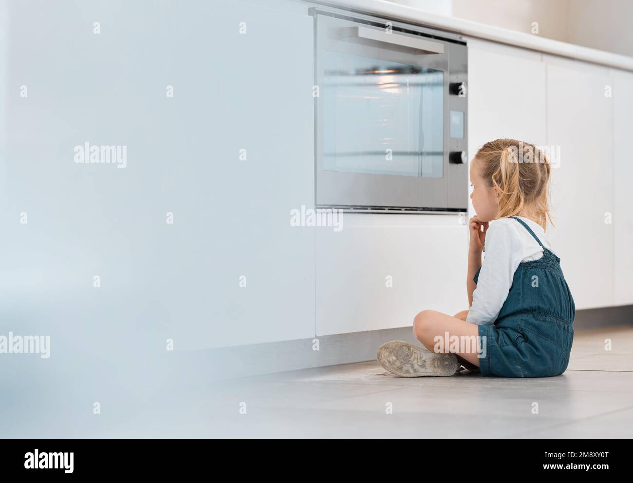 Ein kleines Mädchen, das geduldig am Ofen wartet. Ein weißes Kind, das auf die Ofentür schaut. Das kleine Kind wartet auf seine Backwaren. Blond Stockfoto