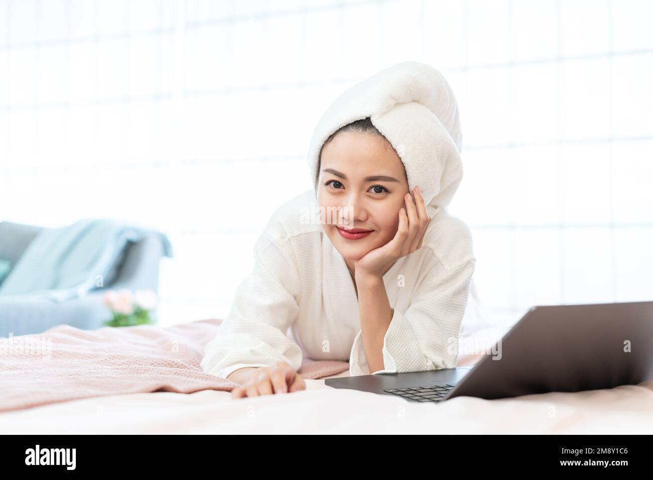 Eine junge Frau, die einen Bademantel trägt, um einen Computer auf dem Bett zu benutzen Stockfoto