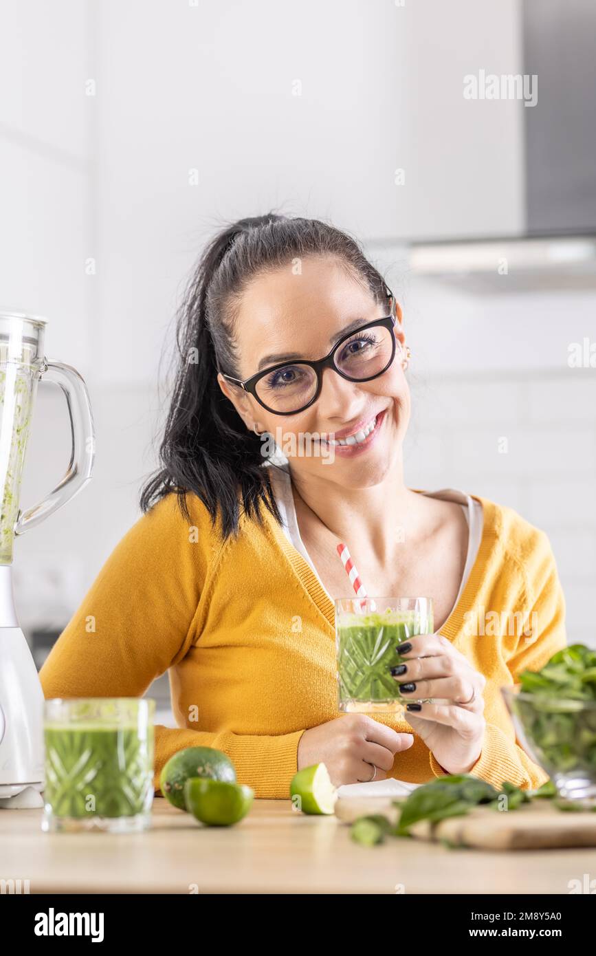 Glückliche Frau mit Spinat-Smoothie-Getränk oder vegetarischem Shake, die in ihrer Küche sitzt. Stockfoto