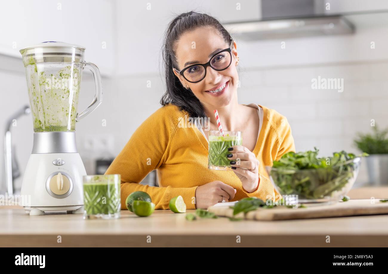 Glückliche Frau mit Spinat-Smoothie-Getränk oder vegetarischem Shake, die in ihrer Küche sitzt. Stockfoto