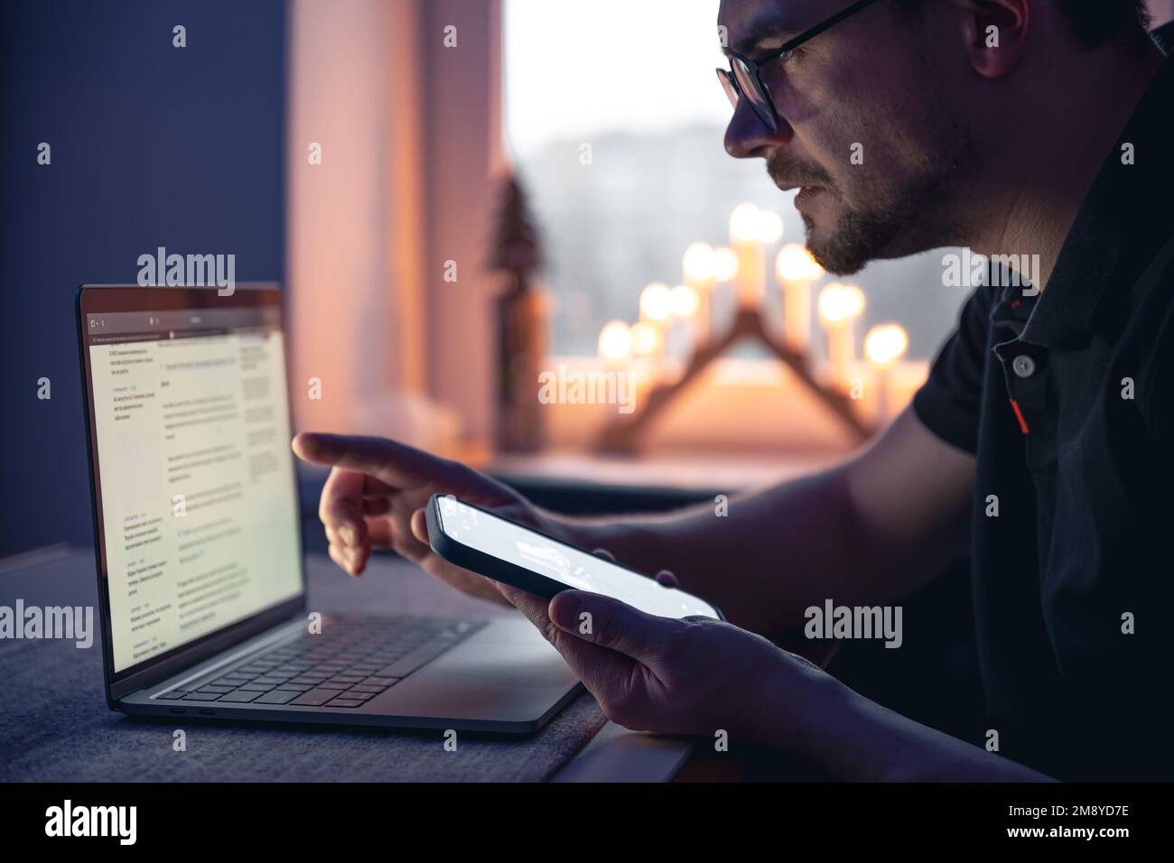 Ein Mann mit einem Smartphone sitzt spät in der Nacht vor einem Laptop. Stockfoto