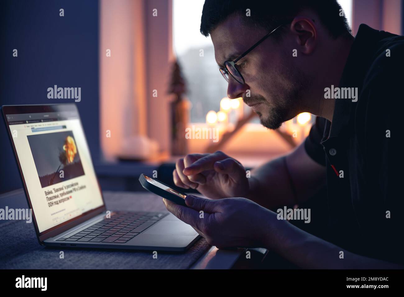 Ein Mann mit einem Smartphone sitzt spät in der Nacht vor einem Laptop. Stockfoto