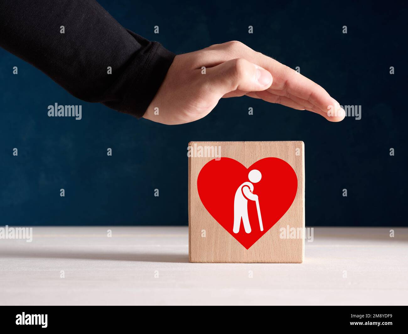 Konzept für Sicherheit, Unterstützung, Versicherung oder Betreuung von Senioren. Männliche Hand steht mit einer Ikone eines älteren Ruheständlers protektiv über dem Holzwürfel. Stockfoto