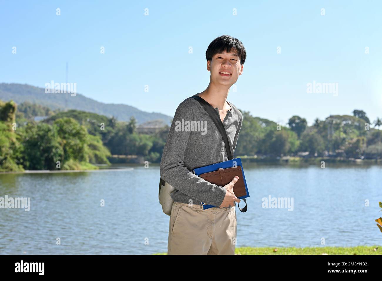 Hübscher junger asiatischer Student in legerer Kleidung, schlendert durch den Campuspark mit einem wunderschönen Teich hinten. University Student Lifestyle CO Stockfoto
