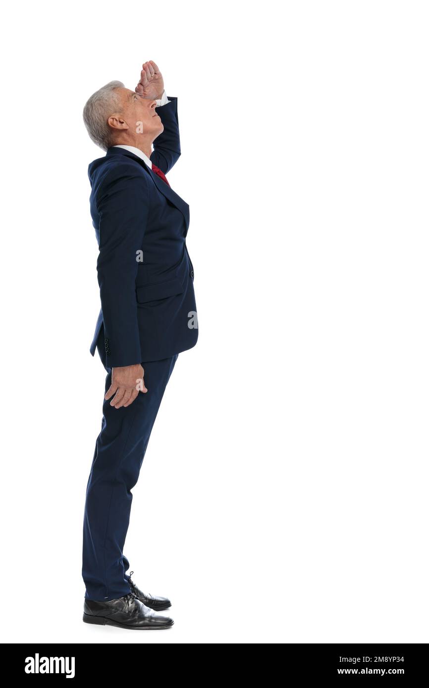 Ganzkörperbild und Seitenansicht eines alten Geschäftsmannes, der seine Augen vor Sonnenlicht schützt, um besser zu sehen, was über ihm ist Stockfoto