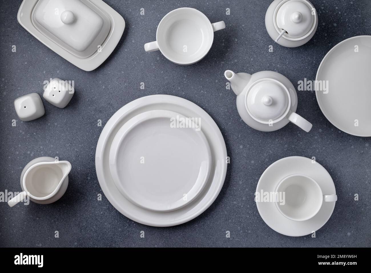 Weißes Geschirr zum Servieren und Essen. Leere saubere Teller, Tasse, Teekanne, Zuckerdose, Milchkanne und Butterschale auf einem grauen Betontisch, Draufsicht Stockfoto