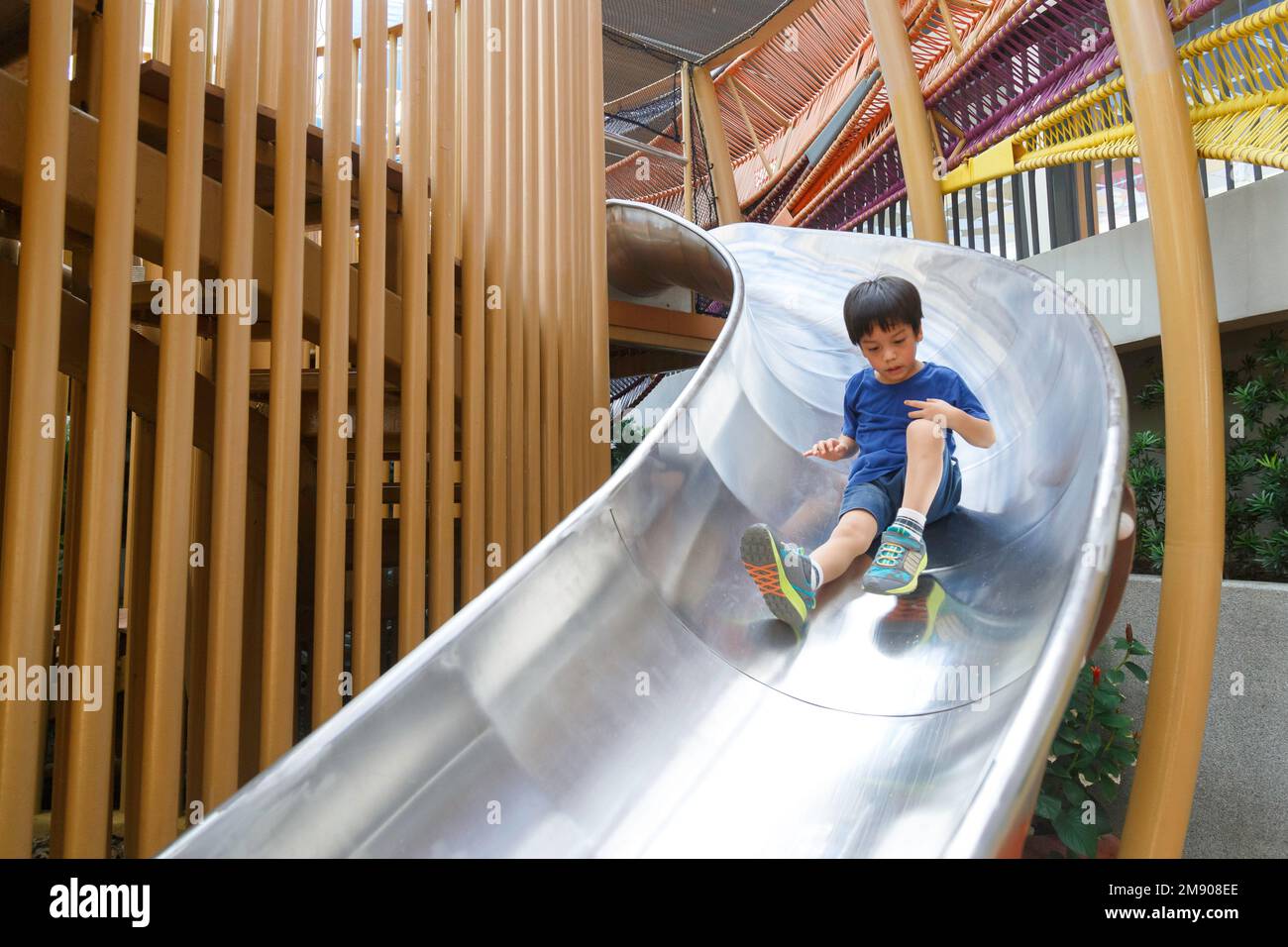 Glücklicher asiatischer Junge auf dem Spielplatz. Das Kind rutscht auf dem Schieber in der Vorschule oder im Kindergarten nach unten. Stockfoto