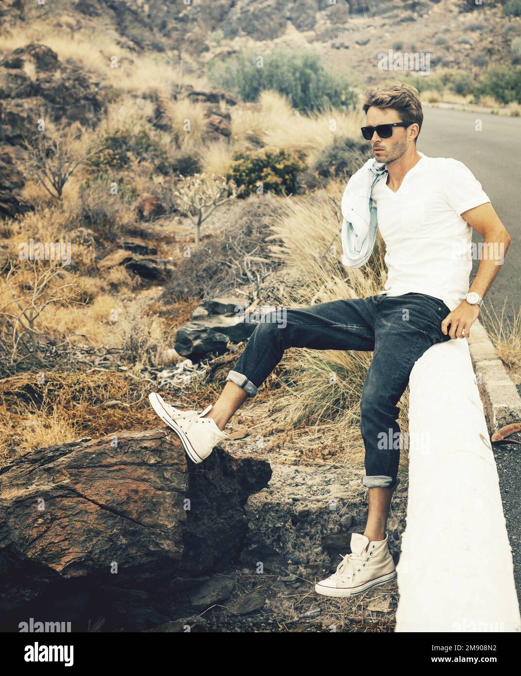 Ein hübscher junger Mann, der auf der Brüstung am Felsen sitzt, mit Brille, weißem T-Shirt und schwarzen Jeans. Stockfoto