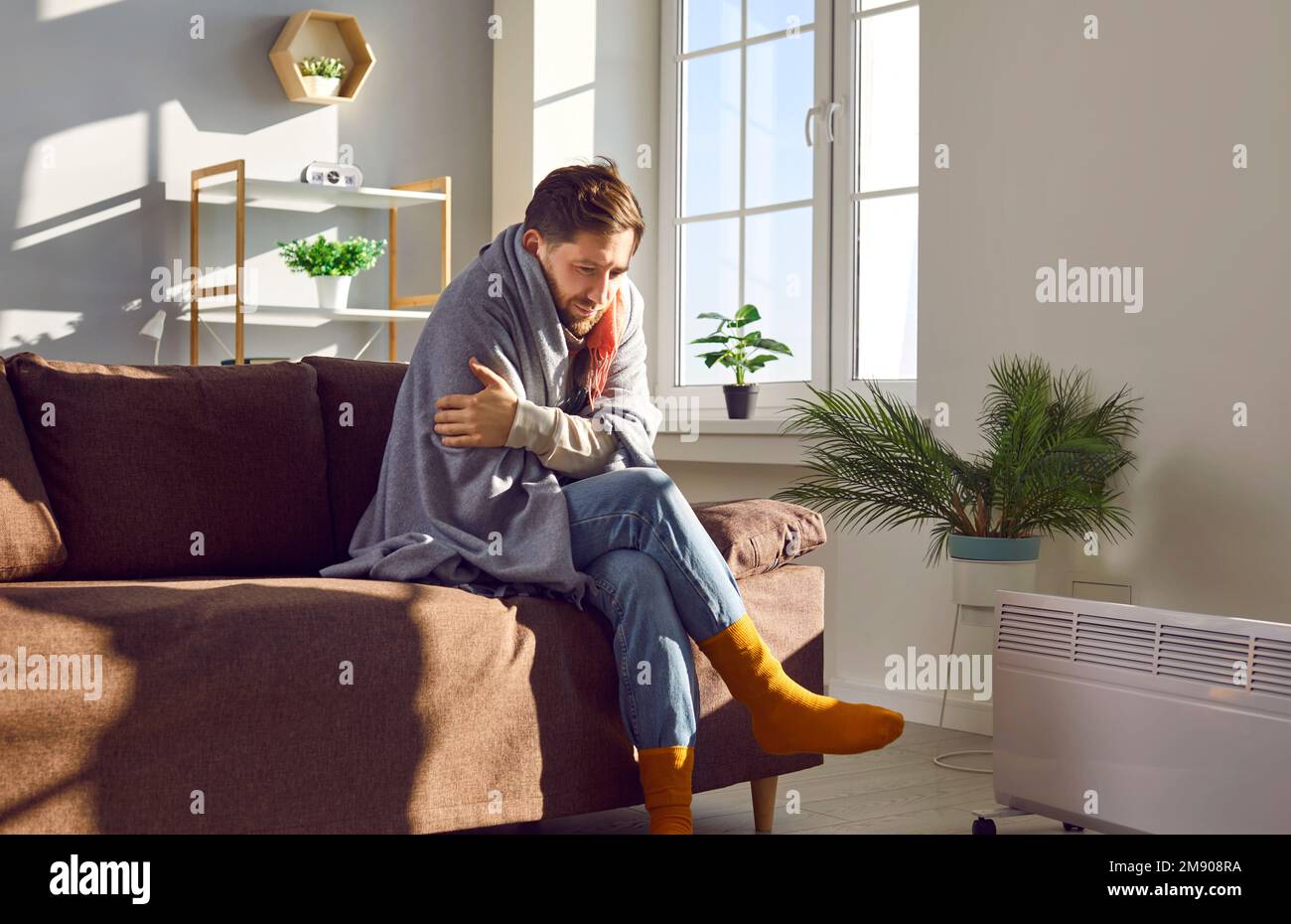 Ein junger Mann mit einer Decke, der auf der Couch neben der Heizung saß und versuchte, sich aufzuwärmen Stockfoto