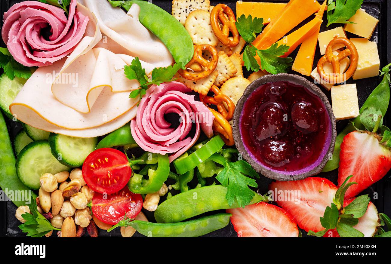Vorspeisenplatte kaltgeschnittener Schinken, Salami, Cracker, Erdbeeren, Gemüse und Käseplatte an Bord vor dunklem Hintergrund. Vorspeisentisch mit Stockfoto