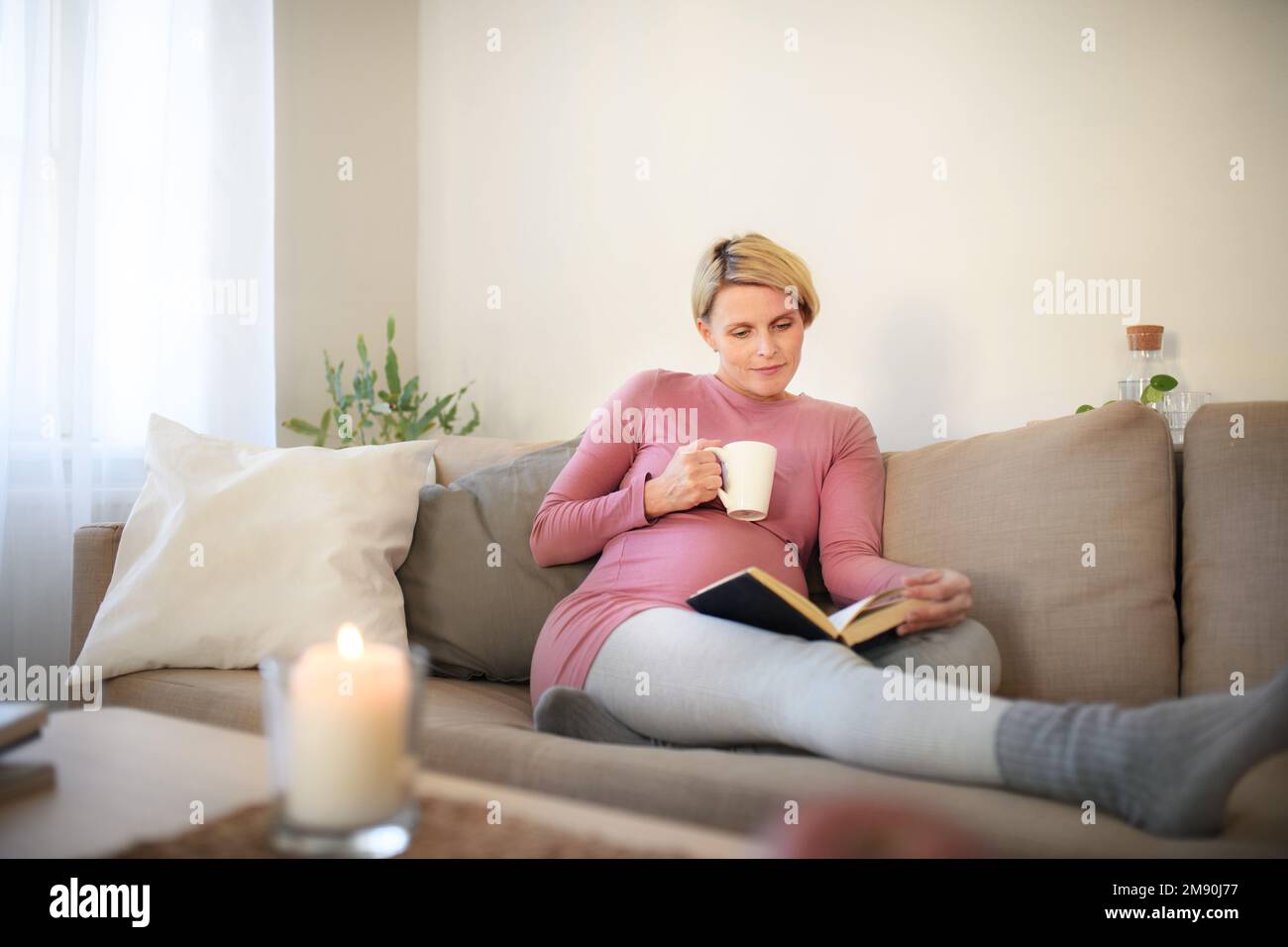 Schwangere Frau, die auf dem Sofa sitzt, Buch liest und eine Tasse Tee genießt. Stockfoto