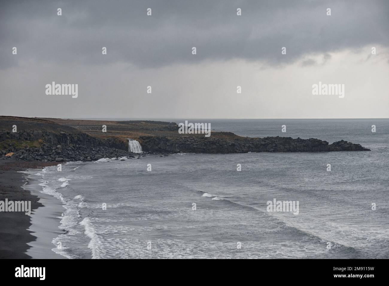 Island Seashore mit schwarzem Sand am Strand und Felsen. Wasserfall im Hintergrund Stockfoto