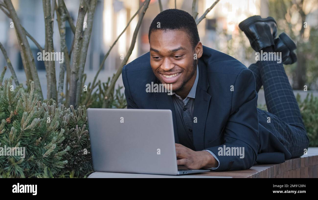 Lächelnder zufriedener, ethnischer Geschäftsmann, Geschäftsangestellter, afroamerikanischer, lustiger Arbeiter, der auf der Werkbank liegt und mit einem Laptop kommuniziert Stockfoto