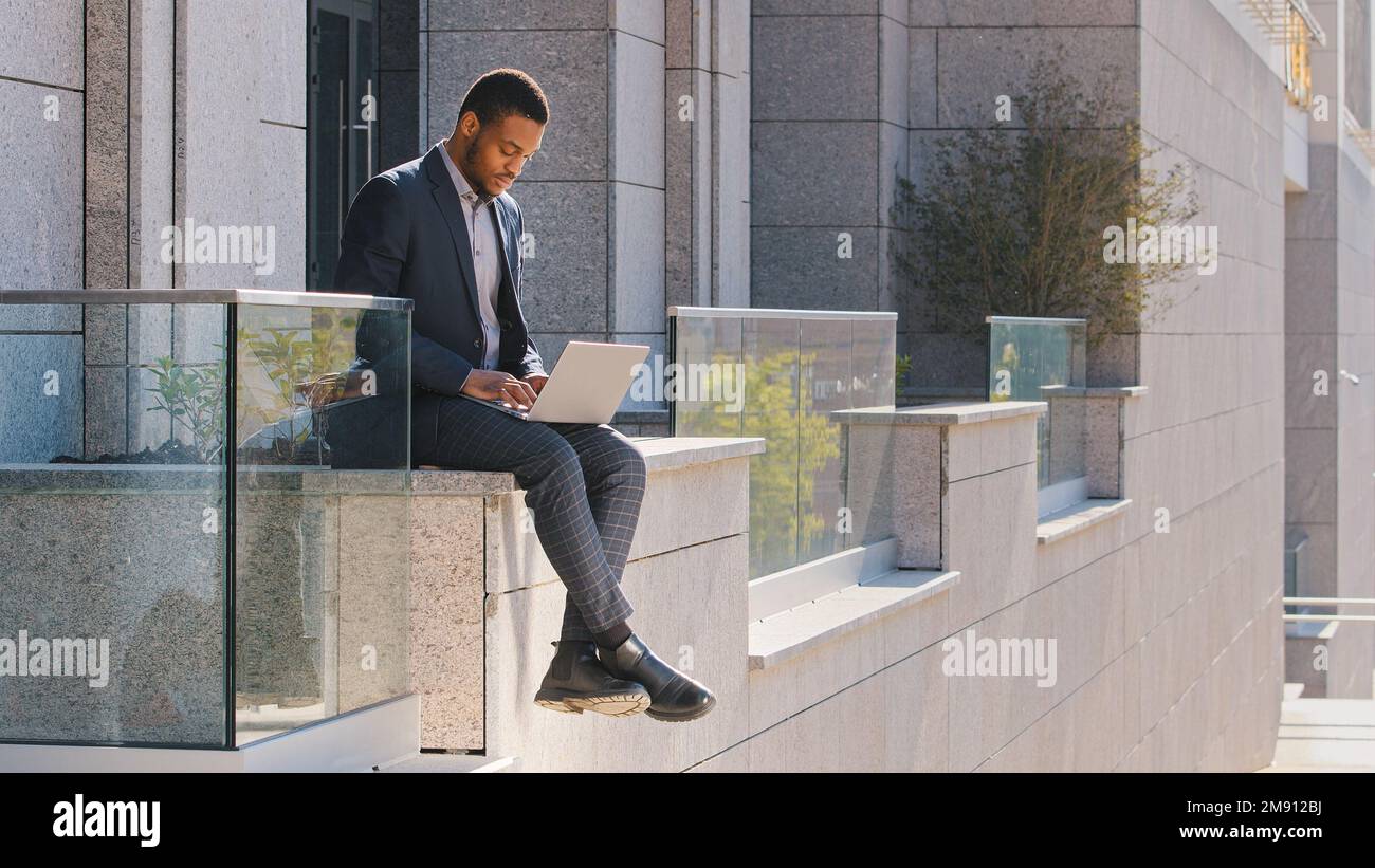 Ethnischer Geschäftsmann, afroamerikanischer Mann, Freiberufler, Unternehmer, Manager, der draußen sitzt und in der Nähe eines männlichen Büroarbeiters auf einem Laptop tippt Stockfoto
