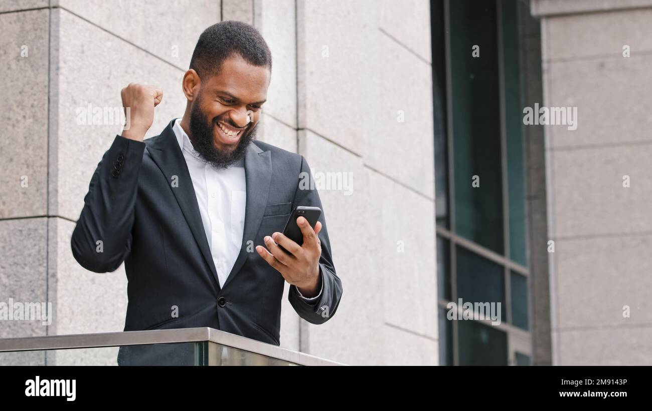 Aufgeregter ethnisch bärtiger afroamerikanischer ethnischer Jahrtausendmann Unternehmer Unternehmer, der auf dem Handy-Bildschirm eine Nachricht liest gute Nachrichten gewinnen Stockfoto