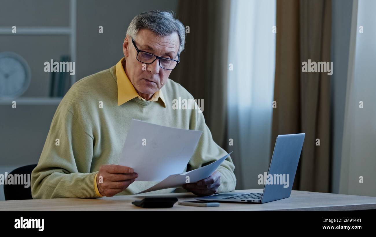 Verwirrter alter weißer, grauhaariger Geschäftsmann, 70s männlicher, trauriger Mann, der mit Papierkram am Tisch sitzt, Probleme mit Dokumenten, Fehler bei der Finanzierung des Budgets Stockfoto