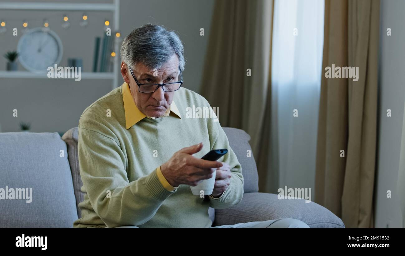 Ein weißer alter alter Mann in einer Brille, der auf der Couch sitzt, den Sender mit Fernbedienung wechselt und Fernsehen ansieht, sich zu Hause entspannt Tee trinkt, Kaffee trinkt, alter Mann Stockfoto
