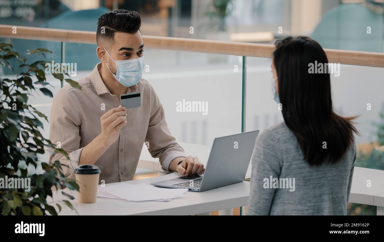 Mann Hispanic Bankangestellter in medizinischer Gesichtsmaske Beratung nicht erkennbare Kundenfrau über Finanzdienstleistungen Debit einfache Zahlung Bankkonto Stockfoto