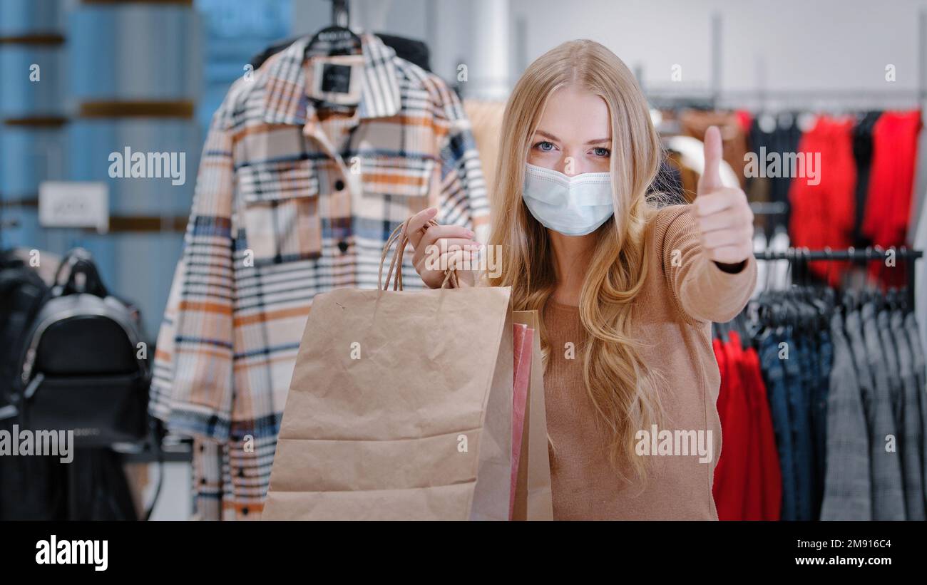 Nahaufnahme glückliche junge Frau in medizinischer Maske während Pandemie Stand in Bekleidungsgeschäft Zulassung Geste Mädchen Verbraucher zeigen Einkaufstaschen zeigen zeigen Stockfoto