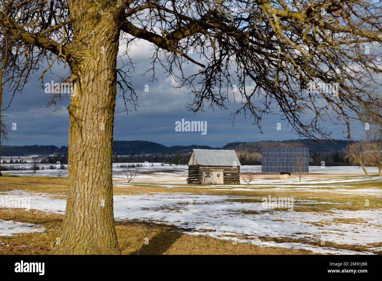 Blockhütte mit Sonnenkollektor im Bauernfeld mit Schneewolken und nackten Bäumen im Winter Stockfoto