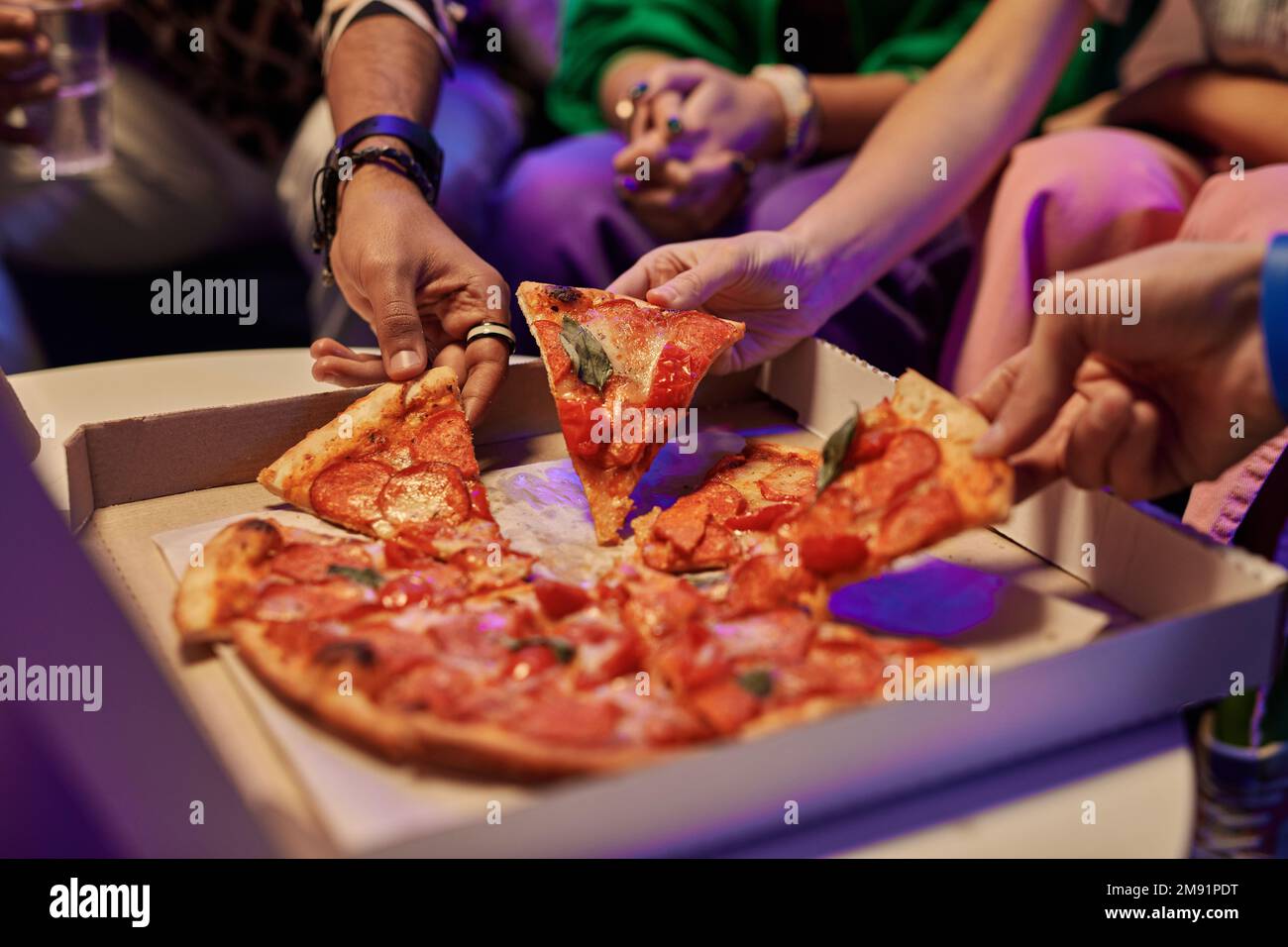 Nahaufnahme von leckeren Pizzaschachteln in quadratischer Pappschachtel und Hände junger Freunde, die sie mitnehmen und essen, während sie die Heimparty genießen Stockfoto