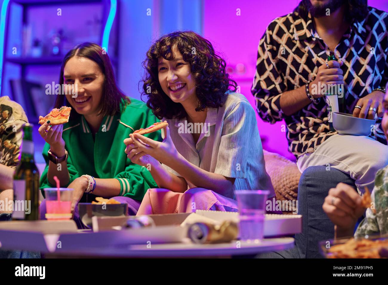 Zwei fröhliche Jugendliche Mädchen in Freizeitkleidung essen Pizza und schauen Comedy im fernsehen, während sie neben einem Kerl sitzen und eine Flasche Bier und Chips trinken Stockfoto