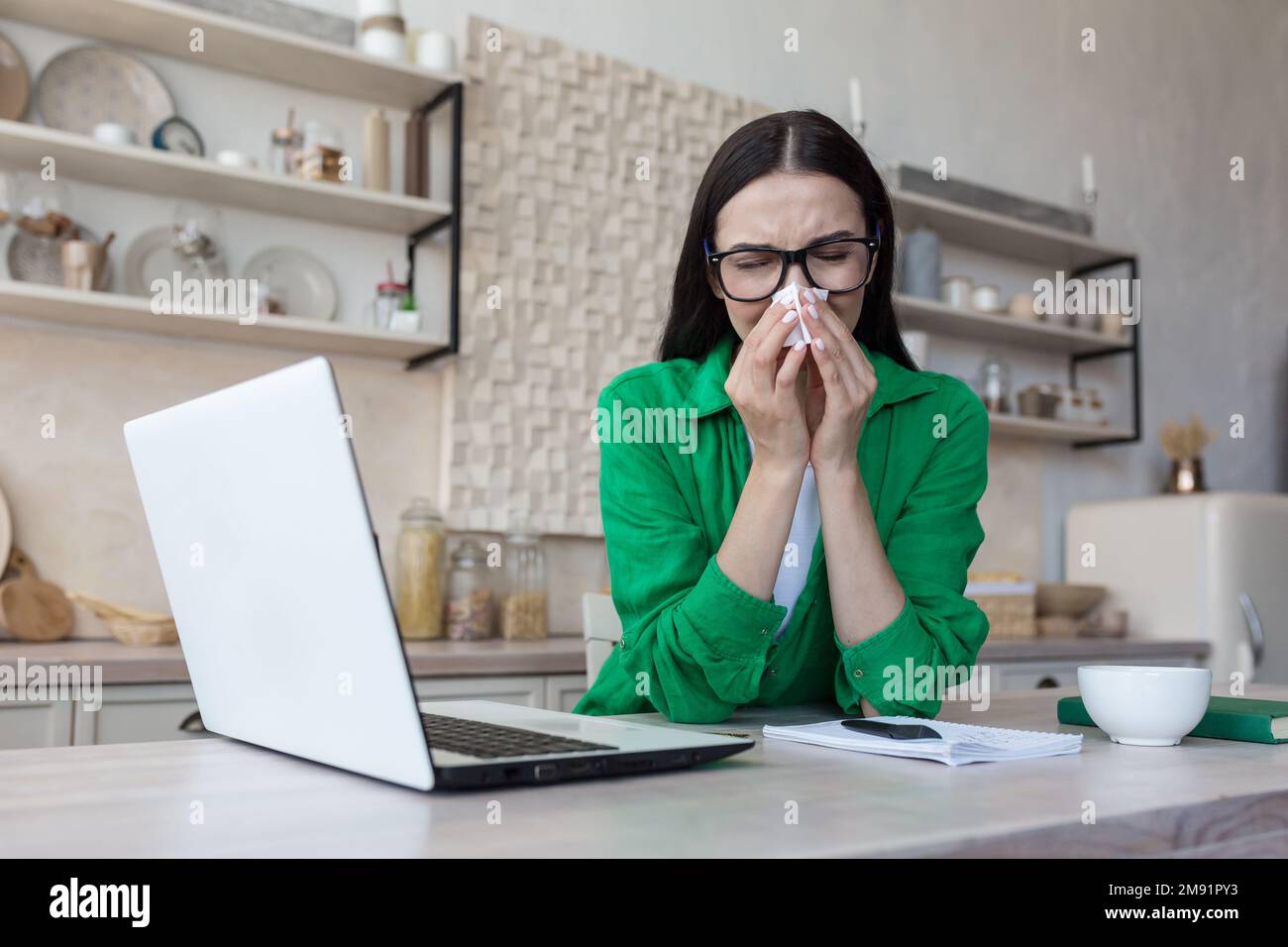 Müde junge Freiberuflerin, die zu Hause in der Küche mit einem Laptop sitzt. Funktioniert. Online-Studien. Krank, fühlt sich schlecht an, wischt die laufende Nase mit einer Serviette ab. Stockfoto