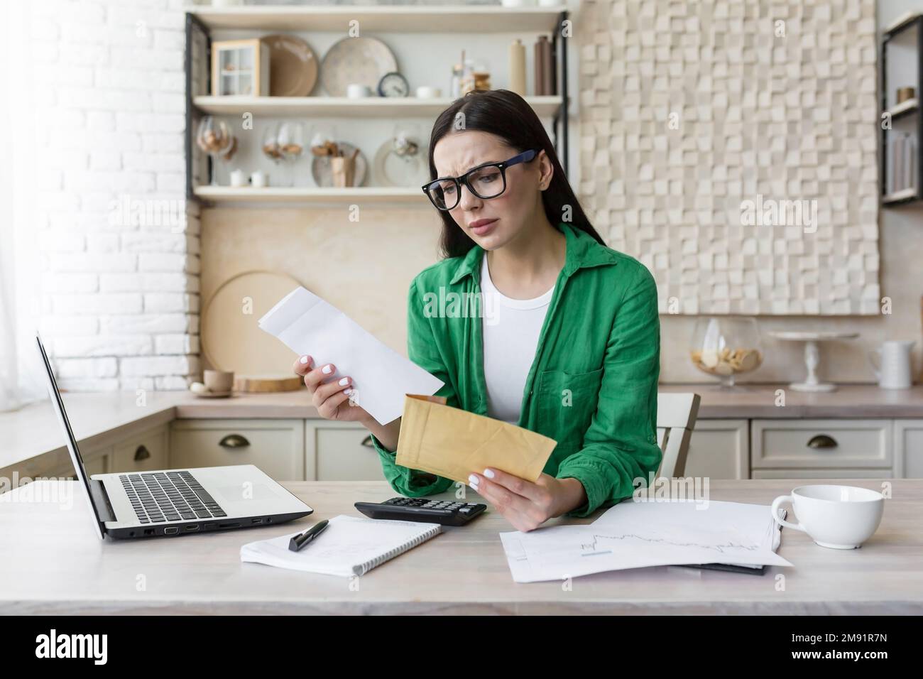 Besorgte junge Frau, die zu Hause in der Küche mit einem Laptop arbeitet. Sie hat einen Umschlag mit einem Brief in der Hand, sie hat schlechte Nachrichten, ein Konto, Schulden, Scheidungspapiere bekommen. Stockfoto