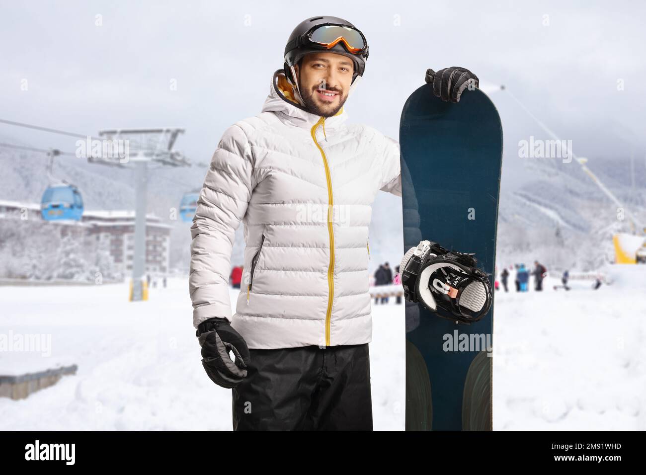 Lächelnder junger Mann mit einem Snowboard, der sich in einem Skigebiet mit einer Seilbahn hinten posiert Stockfoto