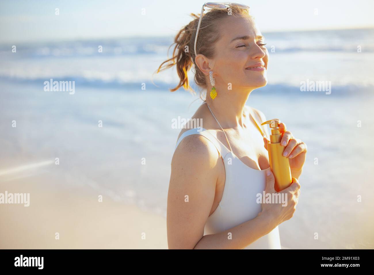 Lächelnde stilvolle 40-jährige Frau in weißem Badeanzug mit Sonnencreme am Strand. Stockfoto