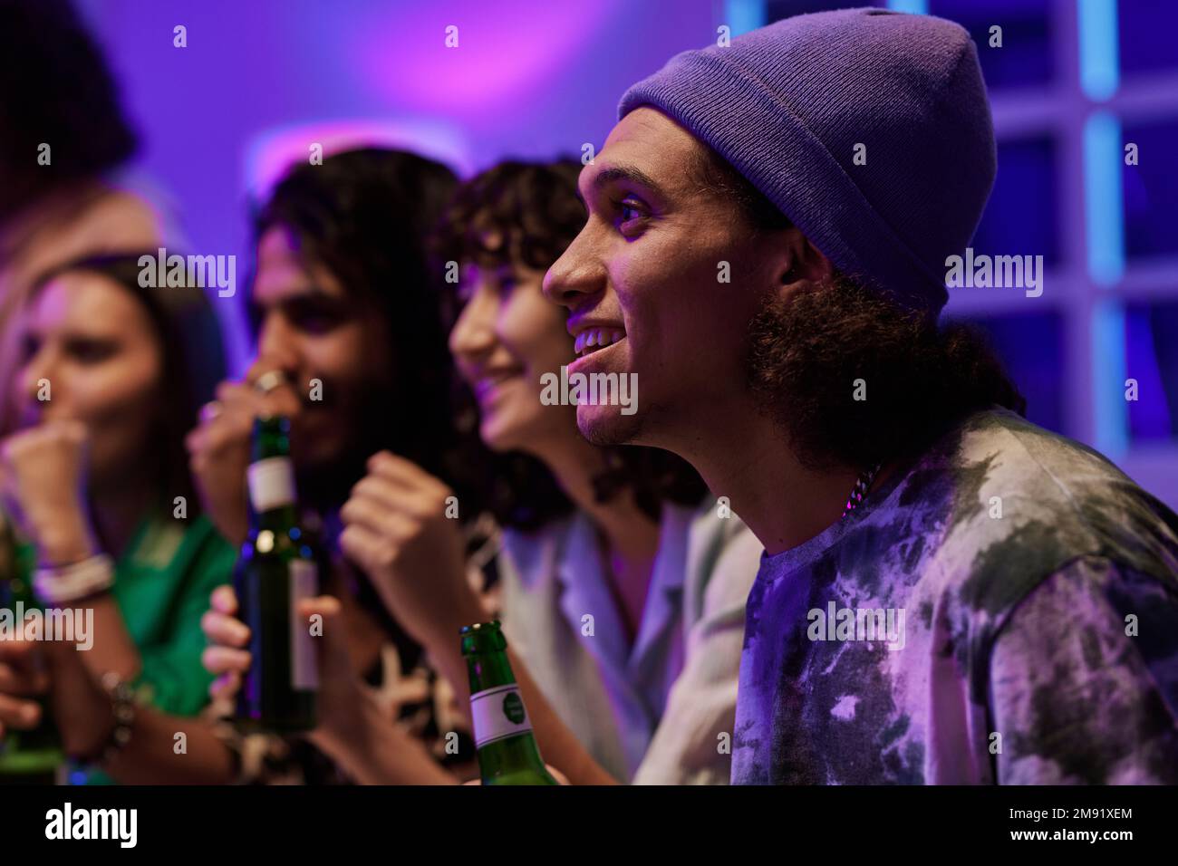 Seitenansicht mehrerer junger interkultureller Freunde, die bei der Versammlung das Spiel ansehen, mit Fokus auf einem glücklichen Kerl mit Beanie-Hut Stockfoto
