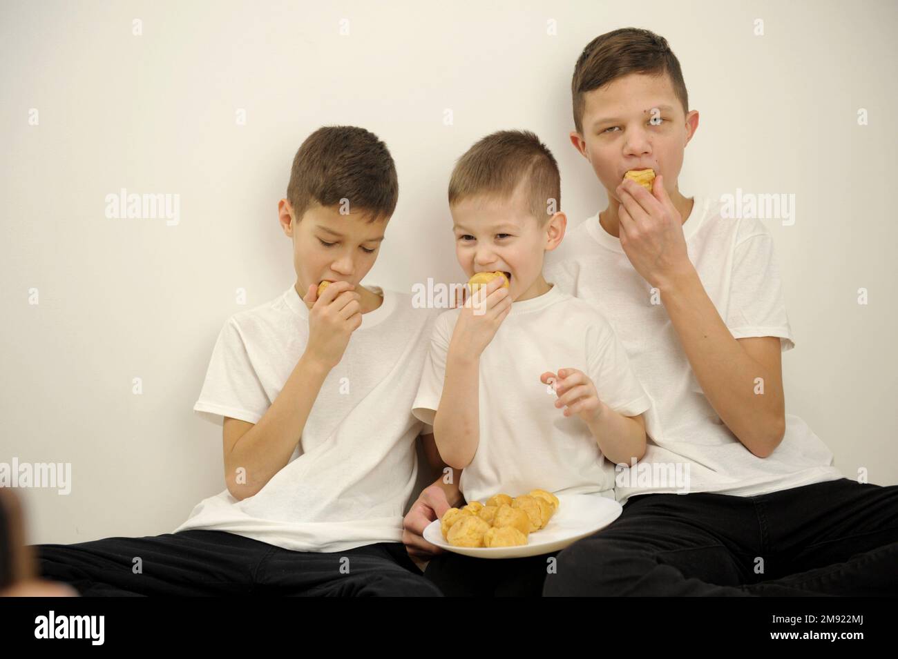 Glückliche, fröhliche Familie, die zusammen Muschis aß, öffneten ihren Mund und beißen köstliche Dessertkinder drei Söhne in weißen T-Shirts drei Kinder Stockfoto
