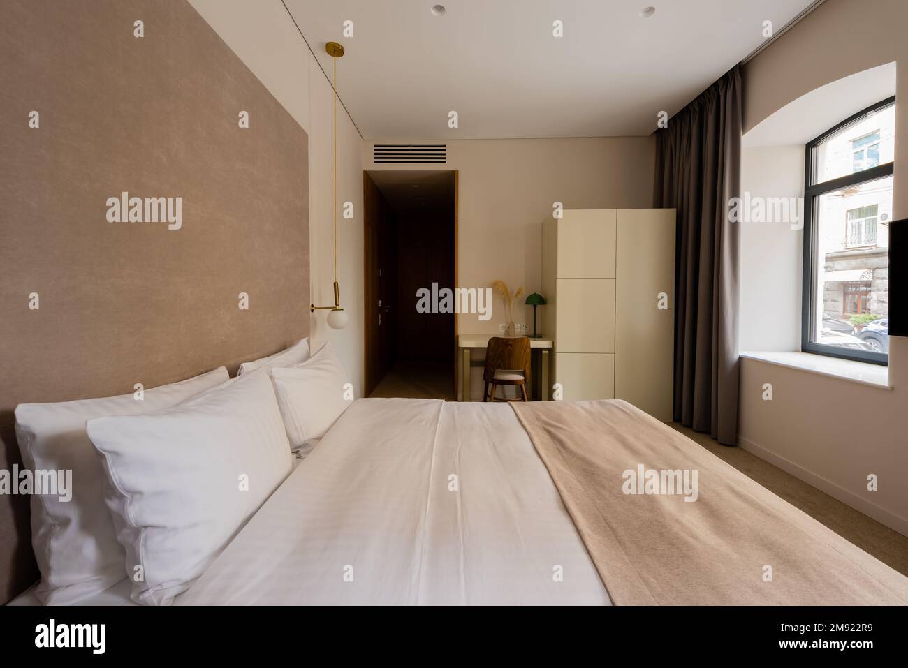Komfortables Bett mit weißer Bettwäsche neben Schreibtisch und Stuhl im modernen Hotelzimmer, Stockbild Stockfoto