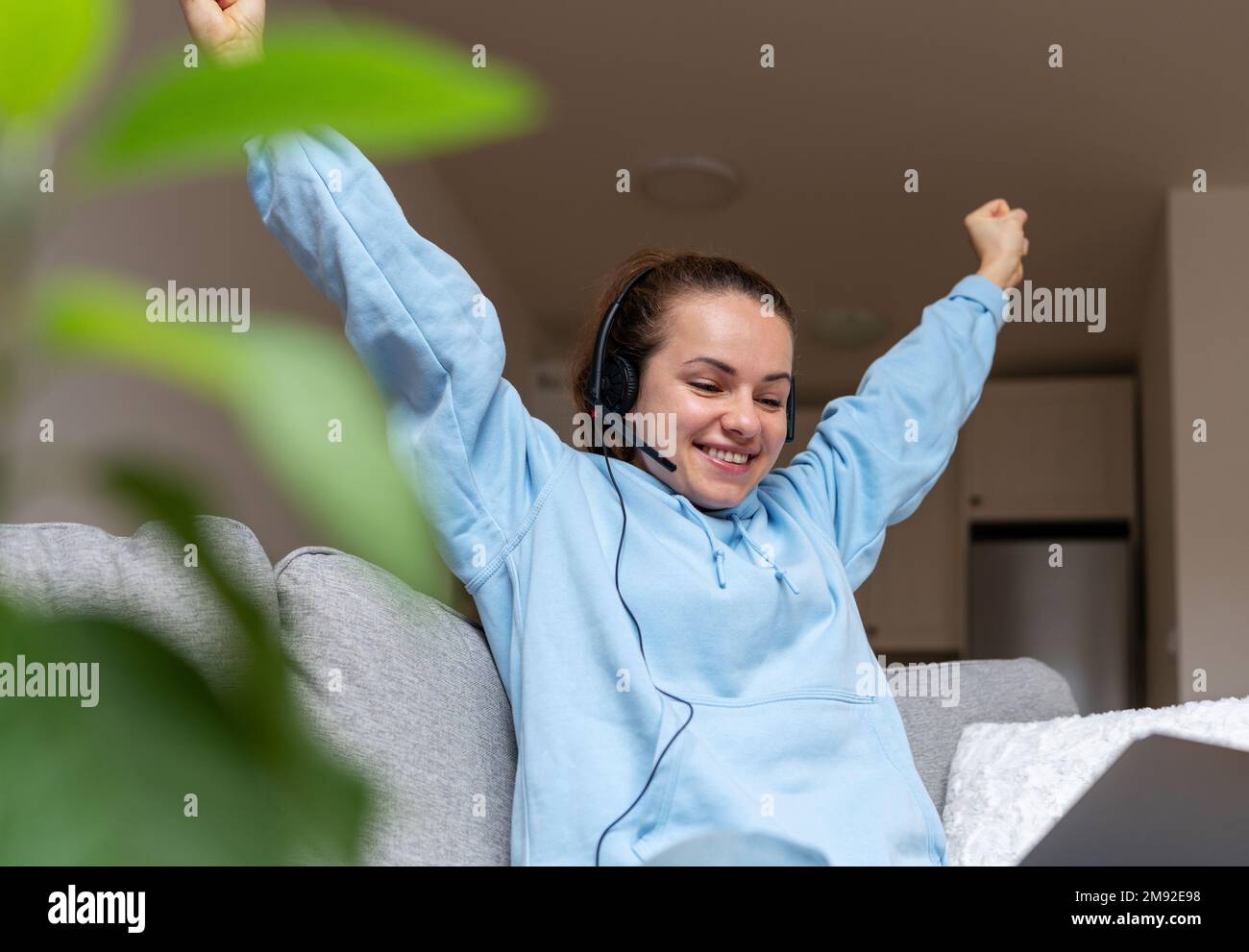 Junge Frau mit Headset und erhobenen Armen, die sich zu Hause auf dem Sofa auf dem Laptop anschaute. Stockfoto