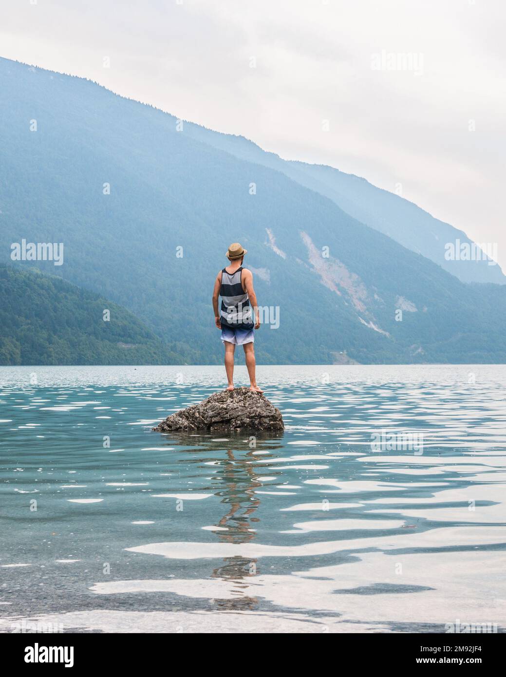 Junger Mann, der auf einem Felsen in der Mitte eines Sees steht, mit Bergen im Hintergrund. Sich jung zu fühlen, die Welt erkunden und bereisen zu wollen. Vertica Stockfoto