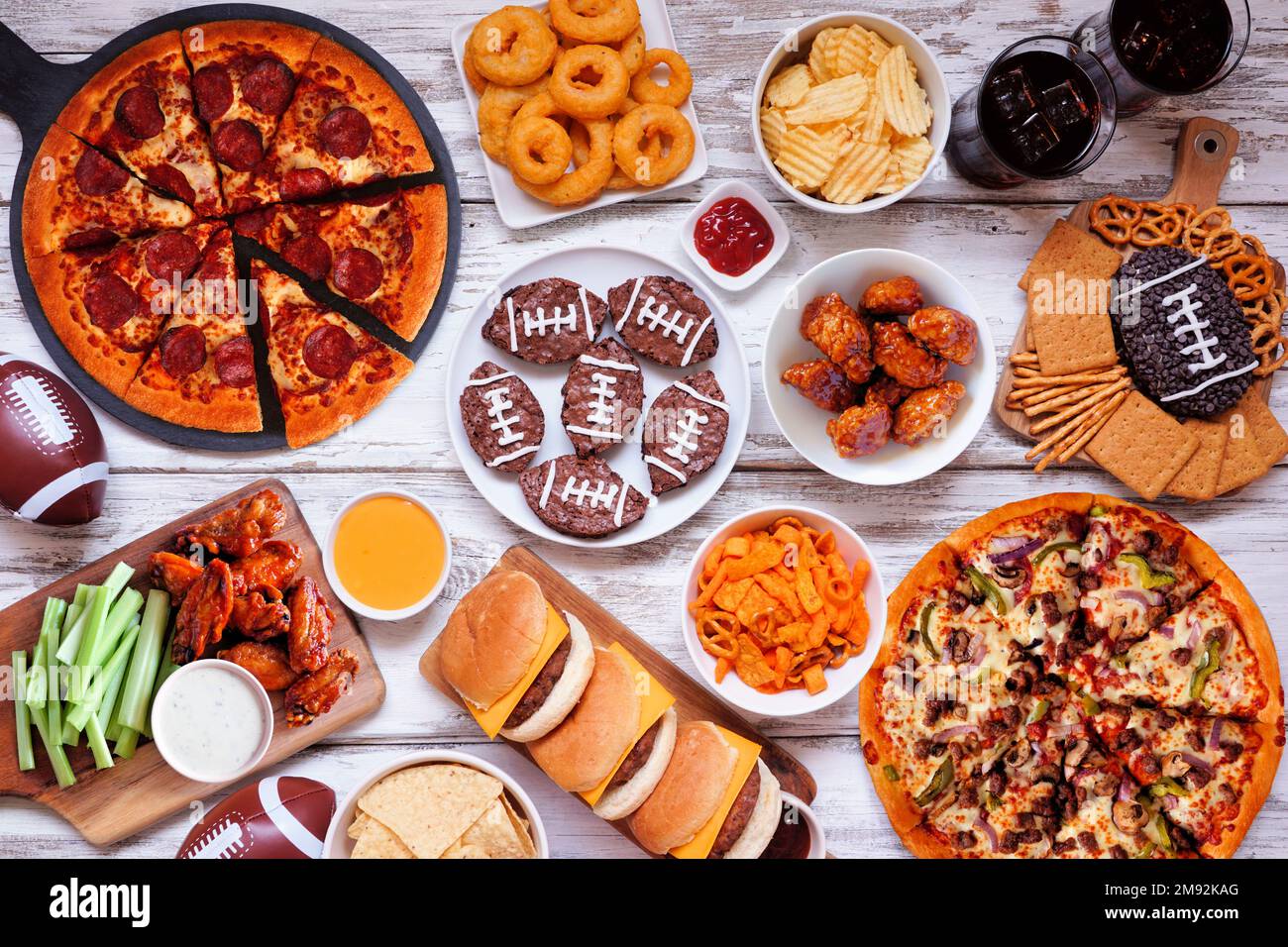 Super Bowl- oder Football-Themen-Esstischszene. Pizza, Hamburger, Flügel, Snacks und Beilagen. Blick von oben auf weißem Holzhintergrund. Stockfoto