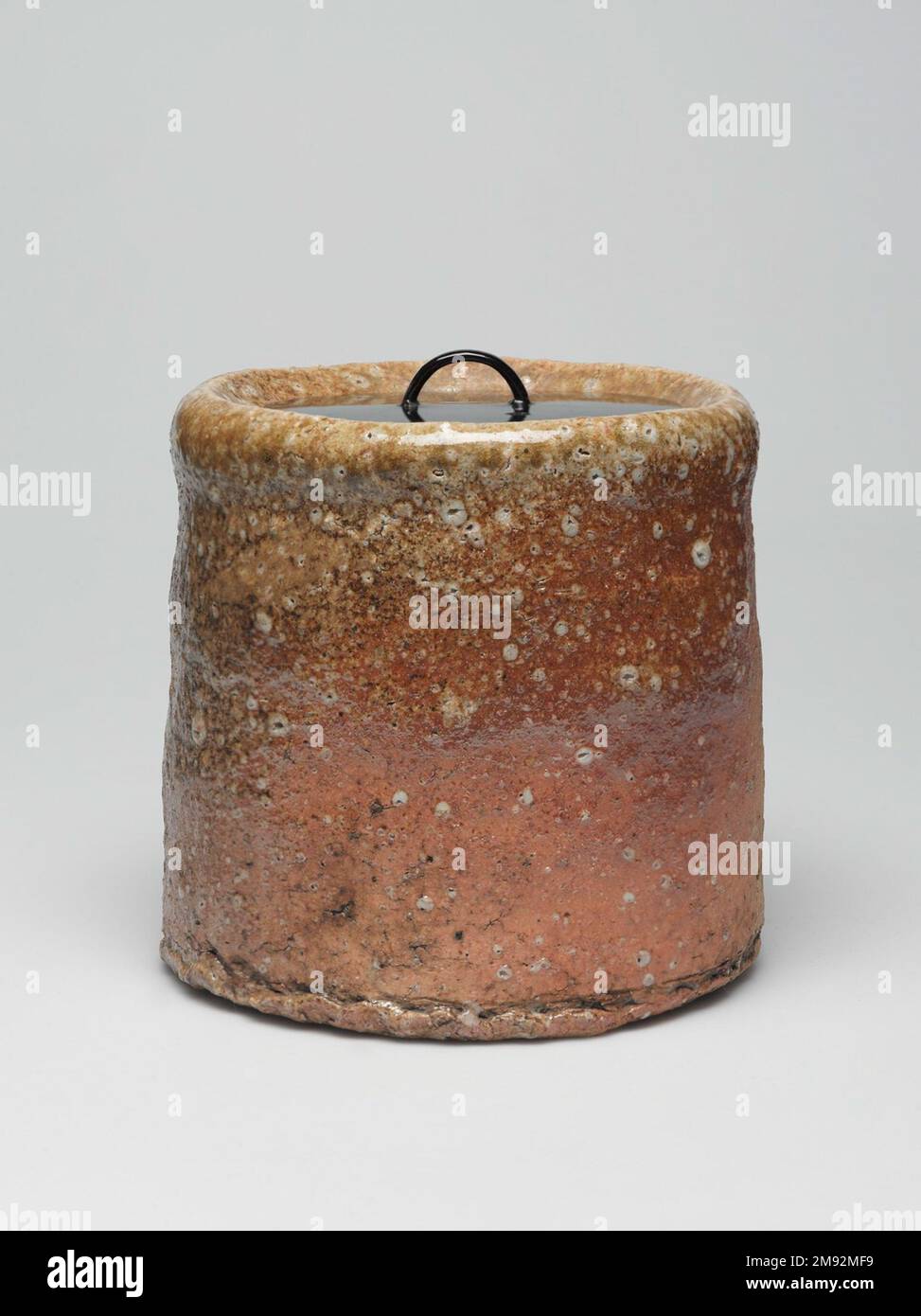 Shigaraki Ware Mizusashi (Teezeremonie Süßwasserdose) Shigaraki Ware Mizusashi (Teezeremonie Süßwasserdose), ca. 1620" Steingut mit Ascheglasur, Lackdeckel; Shigaraki-Geschirr, 6 x 6 1/2" (15,2 x 16,5 cm). Während die meisten Töpfer darauf achten, Steine aus ihrem Lehm zu filtern, verstehen die Töpfer aus der Region Shigaraki, dass die durch steinige Quarzstücke und Feldspat erzeugte Textur ihre Waren taktil und visuell interessant macht. Die Handwerker fügen nur minimale Glasur hinzu, sodass die Oberfläche ihre Körnung behält. Shigaraki, östlich von Kyōto, ist seit Jahrhunderten eine Ofenanlage, die ursprünglich hergestellt wurde Stockfoto