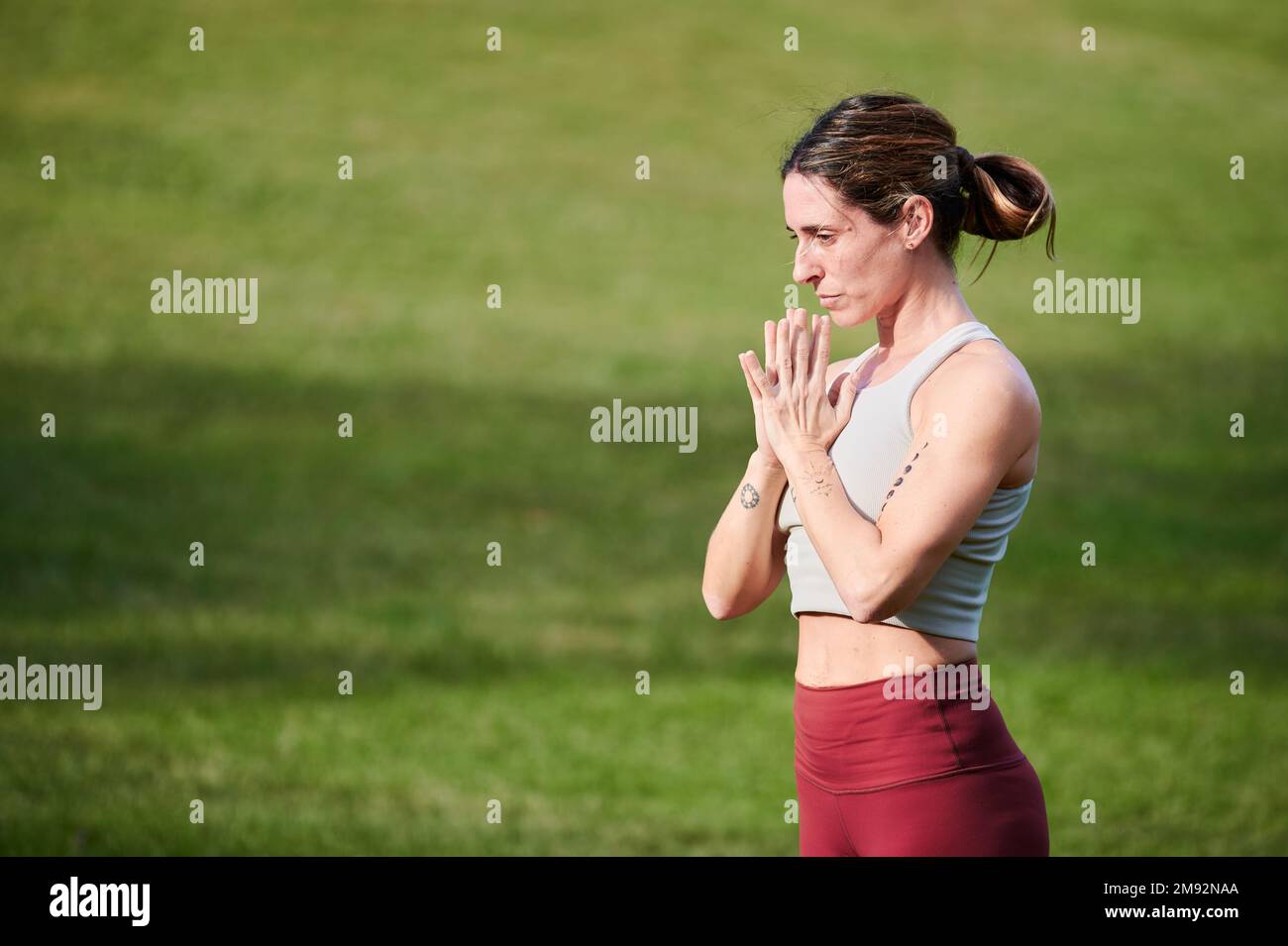 Seitenansicht einer friedlich fokussierten Frau in Sportbekleidung, die wegschaut, mit Gebetshändern meditiert und auf einem grasbedeckten Rasen nachahmt Stockfoto